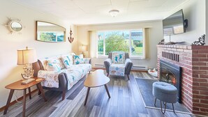 A living room with a sofa, armchair, and ottoman, a fireplace, and a large window with a view of greenery. - PICKLEBALL! Walk to Beach, Fireplace, BBQ (ANC#9) (Long Beach)