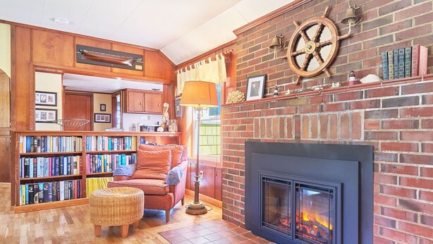 A cozy living room with a fireplace, bookshelves, a comfortable armchair, and a wicker ottoman.