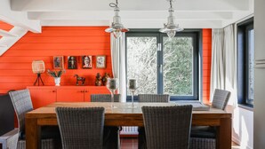 Un coin repas avec une table en bois et des chaises en osier, un mur d’un orange vif et une grande fenêtre avec des rideaux.