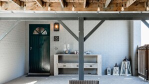 Un espace extérieur abrité, aménagé avec un banc en bois, une chaise en osier et une étagère fixée au mur, ornée d’objets décoratifs.