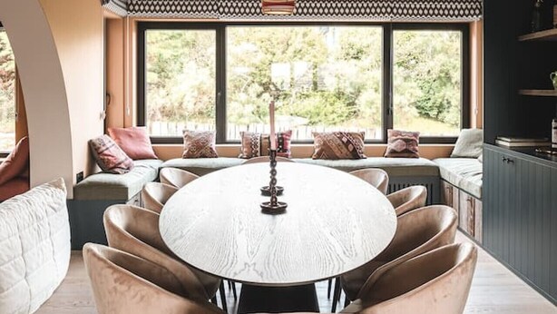 Een moderne eetruimte met een ronde tafel, omringd door comfortabele stoelen, een hanglamp en uitzicht op groen door grote ramen.
