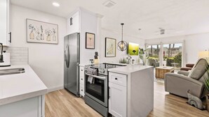 A modern kitchen with white cabinetry, a built-in oven, and a stainless steel refrigerator. There is a dining area with a white table and chairs, and a living room with a grey sofa and a plant.