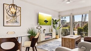 A modern living room with a glass dining table, a sofa, a TV, and a large window with a view of palm trees.