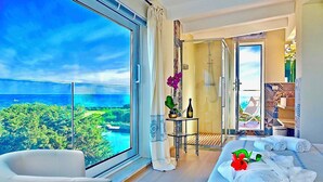 A modern bedroom with a large window overlooking a scenic view, a bed with towels and flowers, a small table with a bottle and flowers, and a shower area. - VILLA DIAMANTE - Seven Bedroom Villa, Sleeps 14 (Stintino)