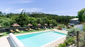 A swimming pool with sun loungers and umbrellas, surrounded by greenery and a forested hillside.