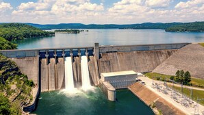 A large dam with multiple water outlets, surrounded by lush greenery and a clear sky.
