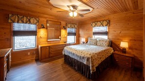 A wooden cabin bedroom with a bed, nightstands, a ceiling fan, and windows with patterned curtains.