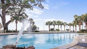 A swimming pool with a water fountain, surrounded by palm trees and lounge chairs.