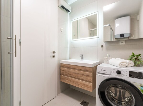 A modern bathroom with a glass shower enclosure, a wooden vanity with a sink, a mirror, a towel dispenser, and a front-loading washing machine. - Modern City Escape (Dubrovnik)