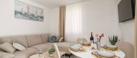 A modern living room with a beige sofa, a dining table set for two, and a painting of sailboats on the wall.