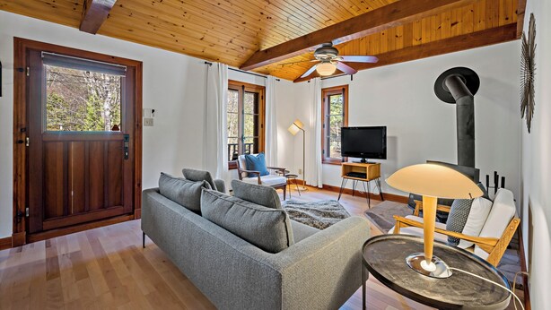A living room with a wooden ceiling, a sofa, a table with a lamp, a TV, and a door with a view of trees.