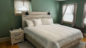 Un dormitorio con cabecera de madera, cama individual, mesita de noche con cajones y ventana con cortinas.