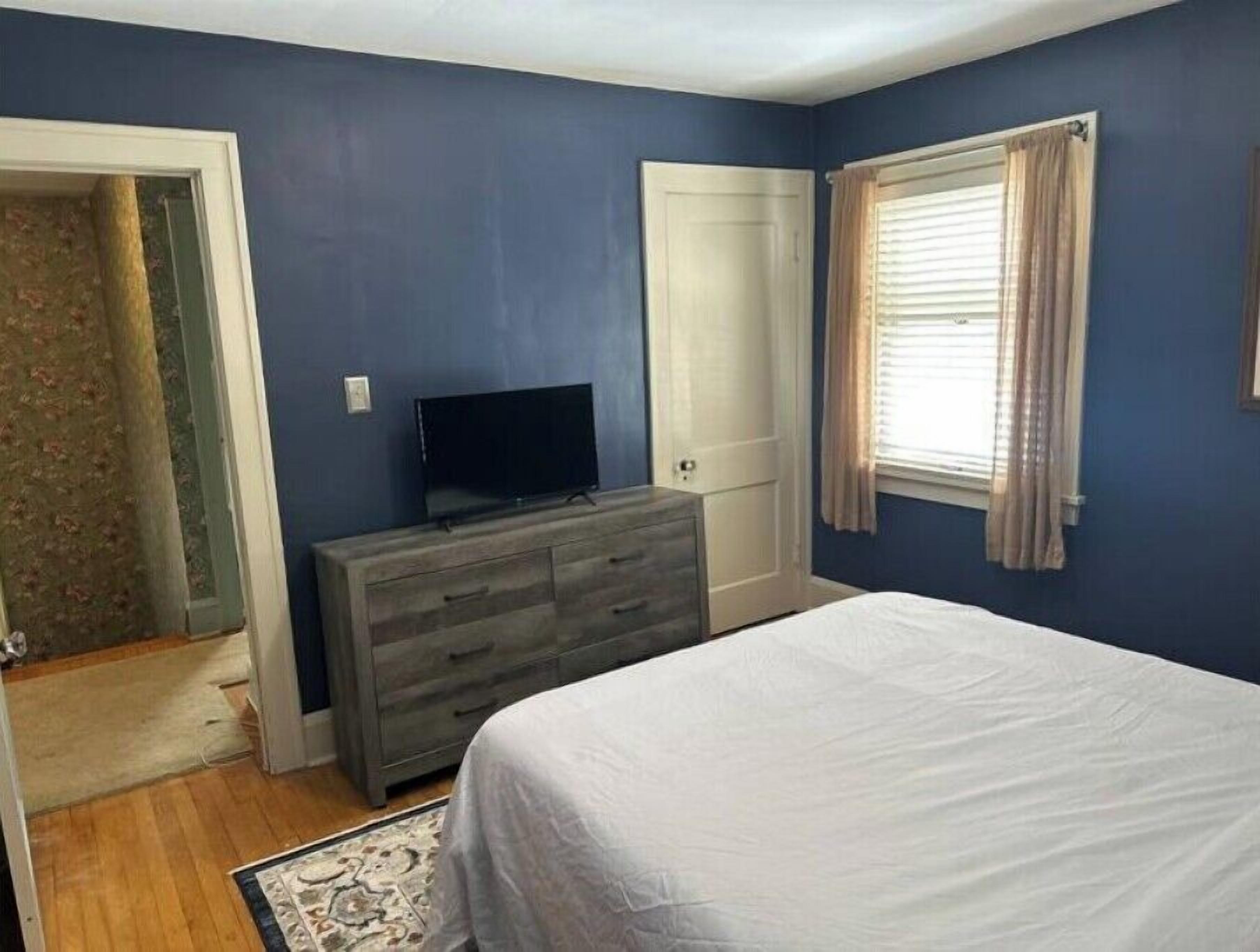 Un dormitorio con cama, cómoda, televisor, puerta y ventana con cortinas.