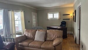 Una sala de estar con un sofá de cuero, una mesa de centro de vidrio, un escritorio y una silla. Hay ventanas con cortinas, una puerta y una lámpara en la pared.