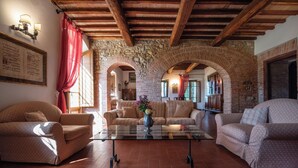 A cozy living room with a stone archway, wooden beams, and a glass coffee table with a vase. - Green Hill Apt In Tuscan Countryside (Siena)