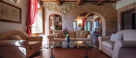 A cozy living room with a stone archway, wooden beams, and a glass coffee table with a vase.