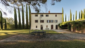 A two-story building with a stone patio, a table with chairs, and tall trees in the background. - Green Hill Apt In Tuscan Countryside (Siena)