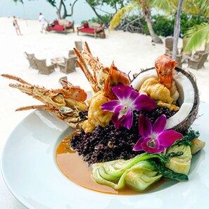 A seafood dish with shrimp, crab, and a coconut shell centerpiece, served on a white plate with a side of greens and a purple flower garnish.