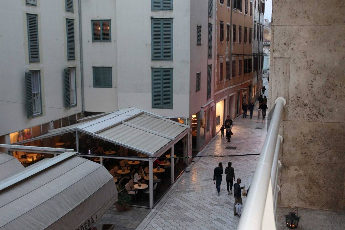 A pedestrian street with outdoor seating, buildings with shutters, and a stone wall.