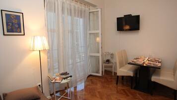 Uma sala de estar com um sofá, uma mesa de jantar posta para dois, uma TV fixada na parede e uma janela com cortinas leves.