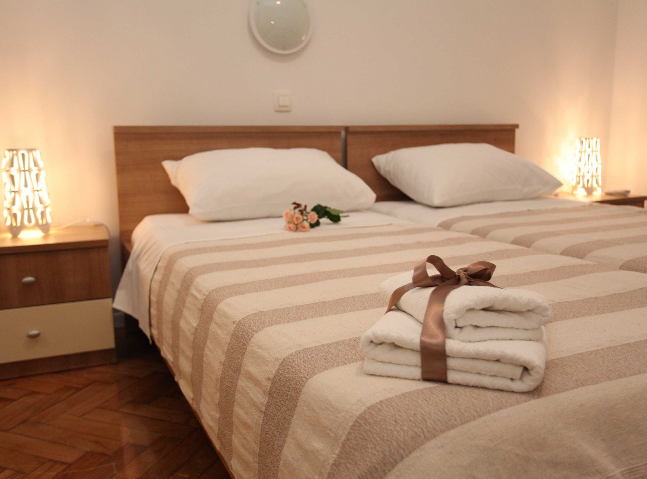 Quarto de hotel com duas camas, cada uma com roupa de cama listrada e cabeceira de madeira. Há duas mesas de cabeceira com luminárias, e um pequeno objeto decorativo em uma cama.