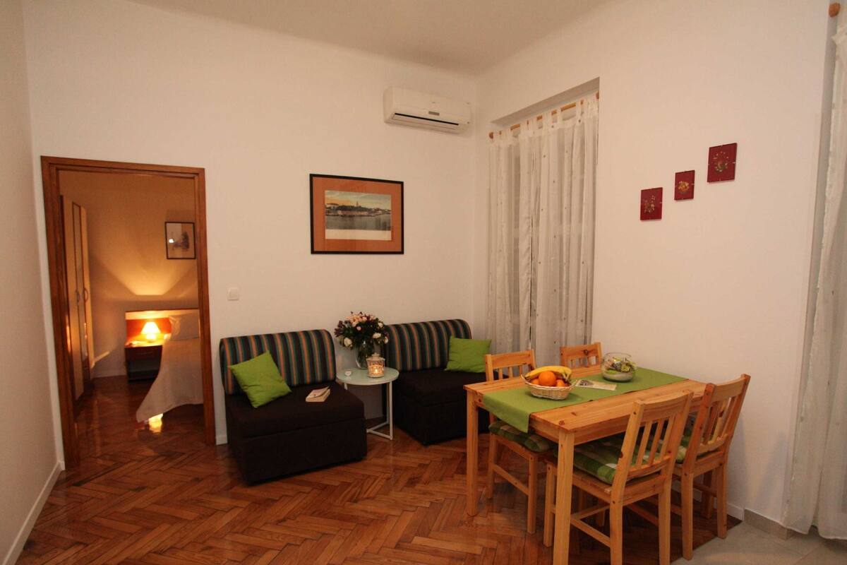 A compact living space with a sofa, a small table, and a dining table with chairs. An air conditioning unit is mounted on the wall, and a framed picture hangs above the sofa.