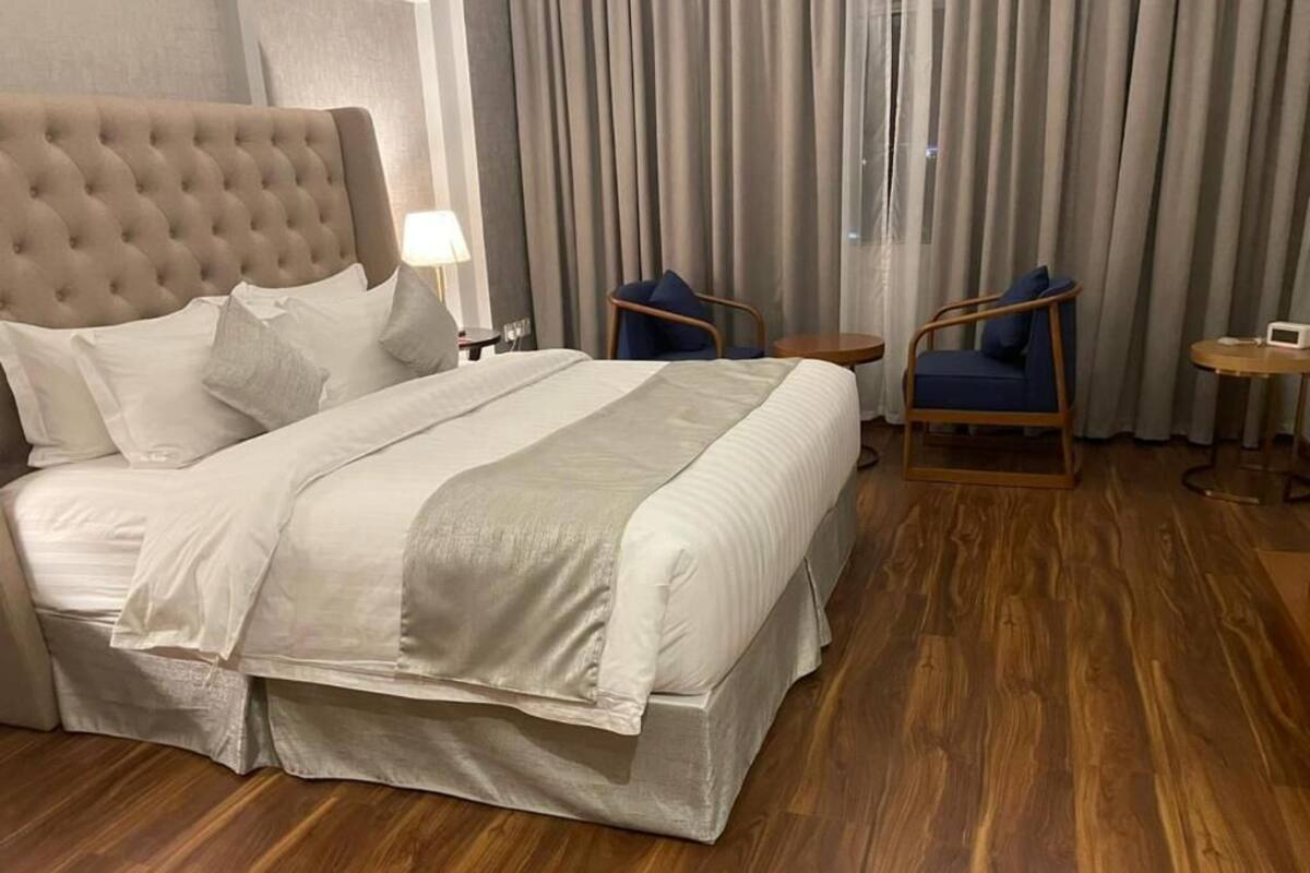 A hotel room with a large bed, wooden flooring, a tufted headboard, two armchairs, a small table, and a lamp.