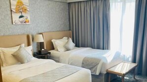 Ein Hotelzimmer mit zwei Betten, einem kleinen Tisch, einer Bank, einer Lampe und einem Gemälde an der Wand.