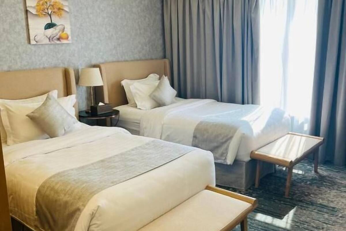 A hotel room with two beds, a small table, a bench, a lamp, and a painting on the wall.
