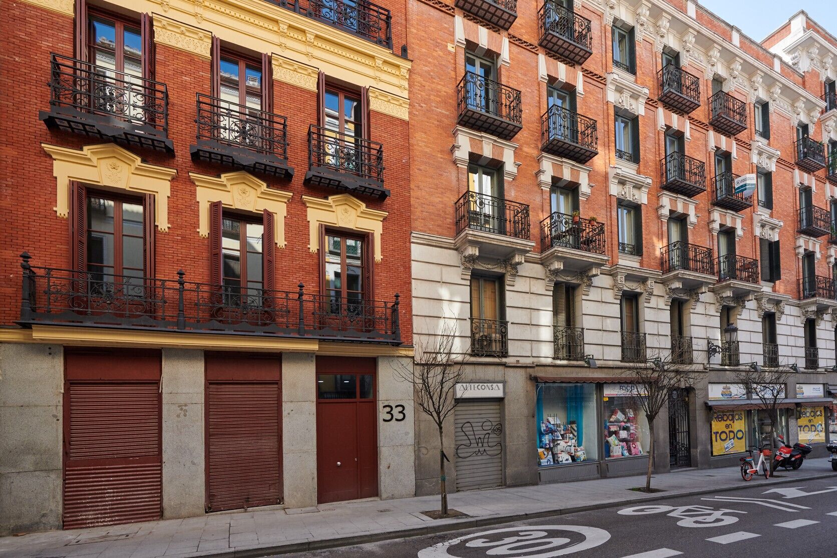 Une rangée d’immeubles en briques rouges, dotés de balcons, portant les numéros 33, et dont les devantures de magasins arborent des enseignes bien visibles.