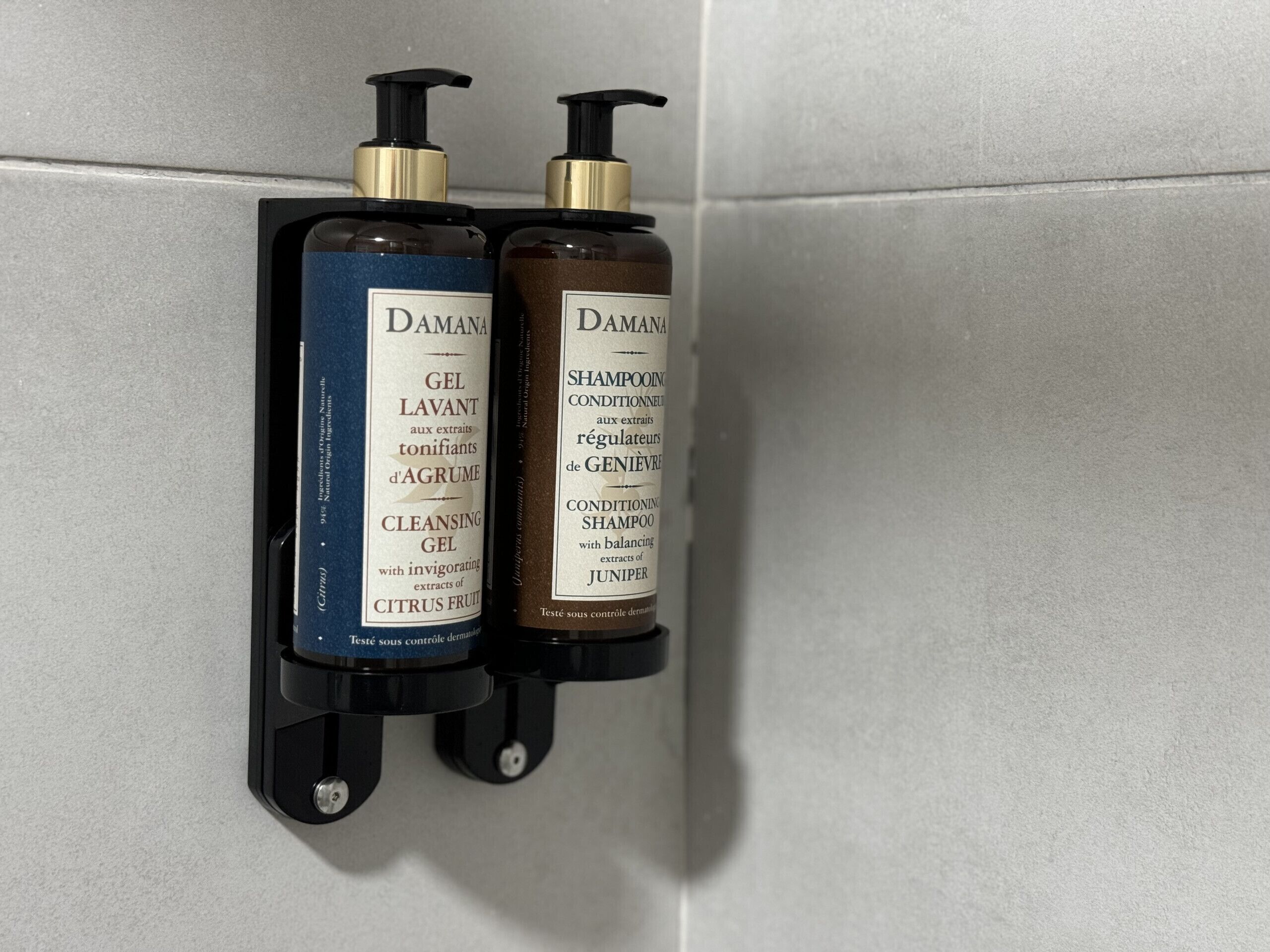 Zwei Flaschen Damana-Produkte, eine als Reinigungsgel und die andere als Conditioner-Shampoo, an einer Wand befestigt.