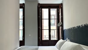 Ein Schlafzimmer mit einem Bett, einem Fenster mit Blick auf Gebäude und einer Tür.