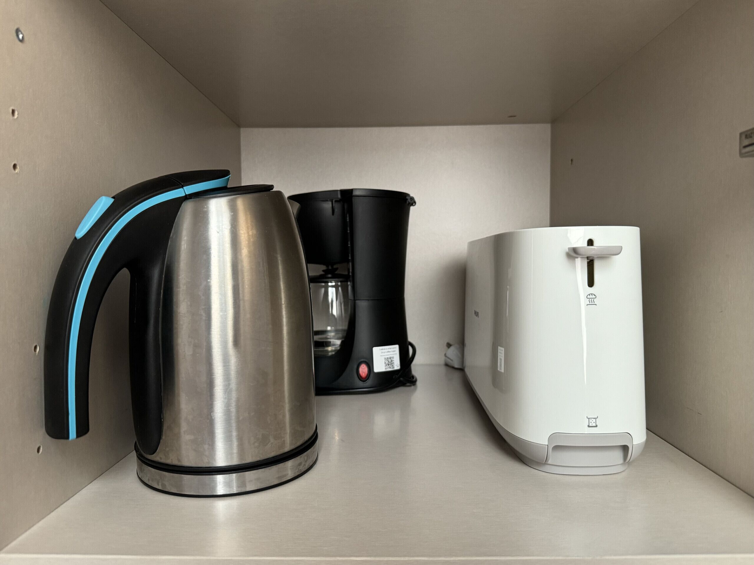 A kitchenette with a stainless steel kettle, a black coffee maker, and a white electric kettle on a countertop.
