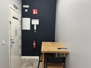 A room with a wooden table, a chair, a fire extinguisher, a wall-mounted phone, and a door with a sign indicating an exit.