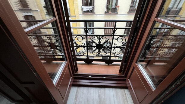 Un balcón abierto con barandilla de hierro forjado, vistas a edificios cercanos y suelo de baldosas.