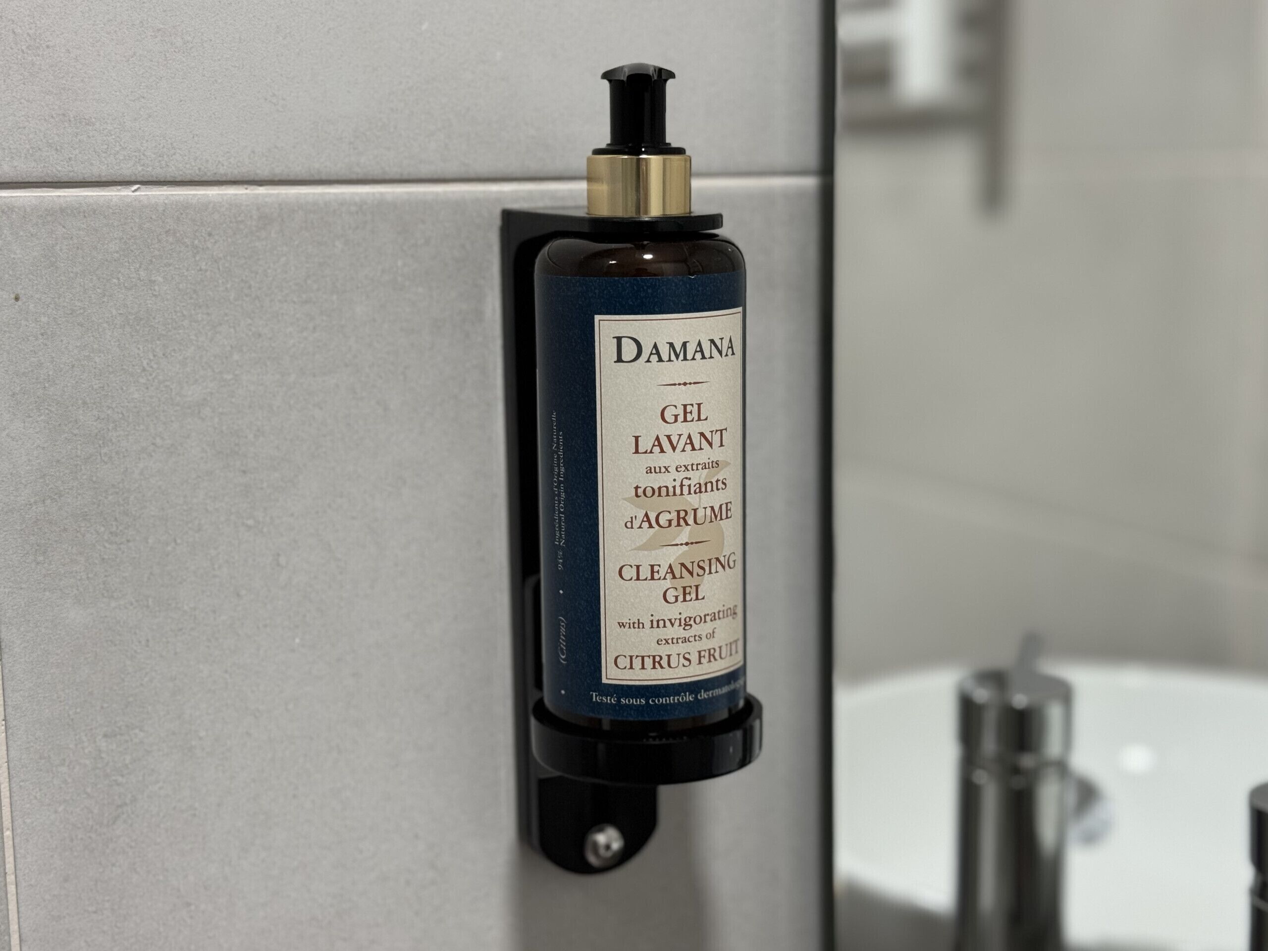 A bottle of Damana cleansing gel with a pump dispenser mounted on a tiled wall.
