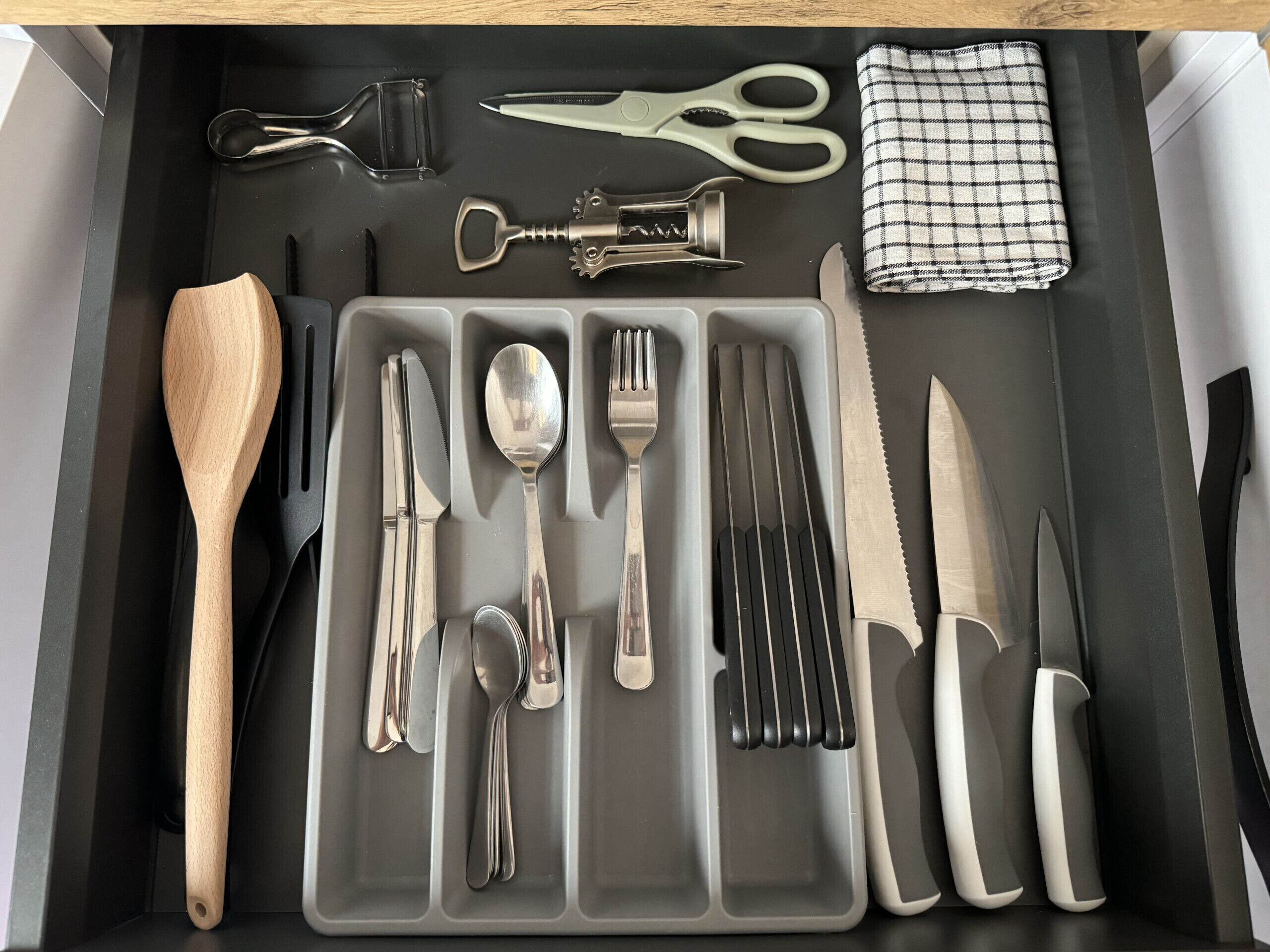 Un tiroir contenant des ustensiles de cuisine bien rangés, notamment une cuillère en bois, des couteaux, des fourchettes, des cuillères et un décapsuleur.