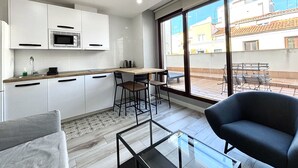 Une cuisine moderne avec des armoires blanches, un plan de travail en bois et un ventilateur de plafond. On y trouve un coin repas avec une table et des chaises, ainsi qu’une grande fenêtre donnant sur les bâtiments environnants.