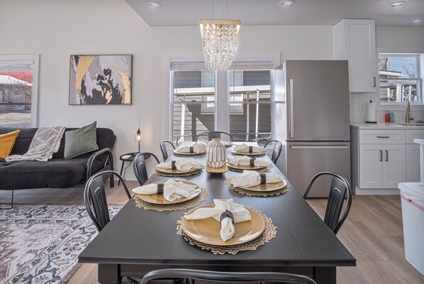 A modern dining area with a dark wooden table set for six, surrounded by black chairs. A chandelier hangs above, and a large abstract painting is on the wall.