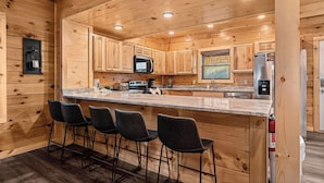A wooden cabin interior with a kitchen featuring a bar area, bar stools, a microwave, a refrigerator, and a sink. - Welcome to Happy Holler Hideaway (Pigeon Forge)