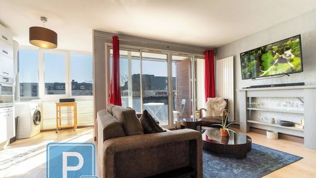 A modern living room with a sofa, armchair, and coffee table, large windows with red curtains, a TV mounted on the wall, and a view of the city.