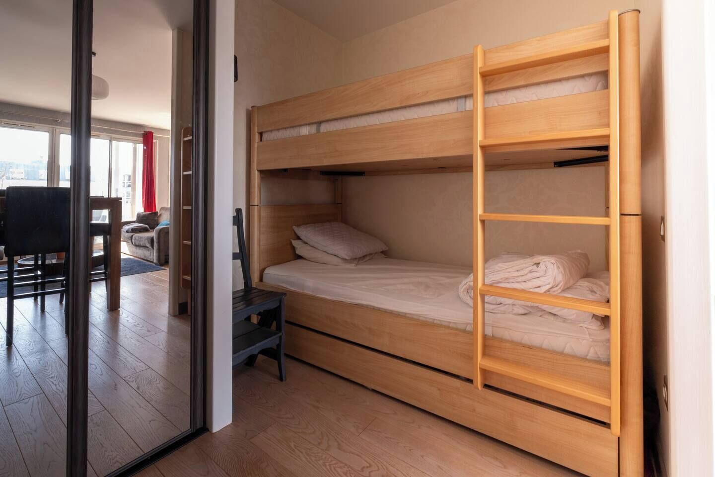 Una cama litera con una escalera, una mesita pequeña y una silla en una habitación con puerta de cristal que da a una sala de estar.