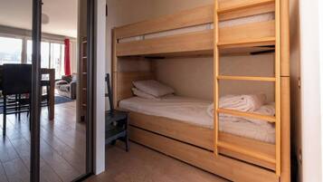 Una cama litera con una escalera, una mesita pequeña y una silla en una habitación con puerta de cristal que da a una sala de estar.