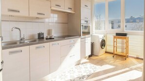 A modern kitchen with light-colored cabinets, a stainless steel sink, and a built-in oven. There's a window providing natural light and a view of buildings outside.