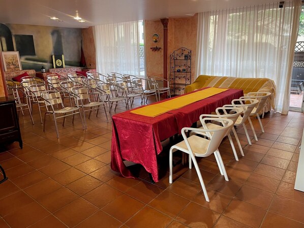 A dining area with a long table covered in a red tablecloth, surrounded by white chairs.