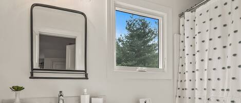 Un baño con tocador de madera, un lavamanos blanco, un espejo, un inodoro y una ventana con vistas a árboles.