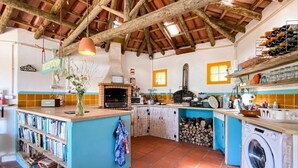 A rustic kitchen with a wooden ceiling, a built-in oven, a washing machine, and a bookshelf filled with books. - Quinta Laranja - Studio with terrace - "Palheiro" (Alvorninha)
