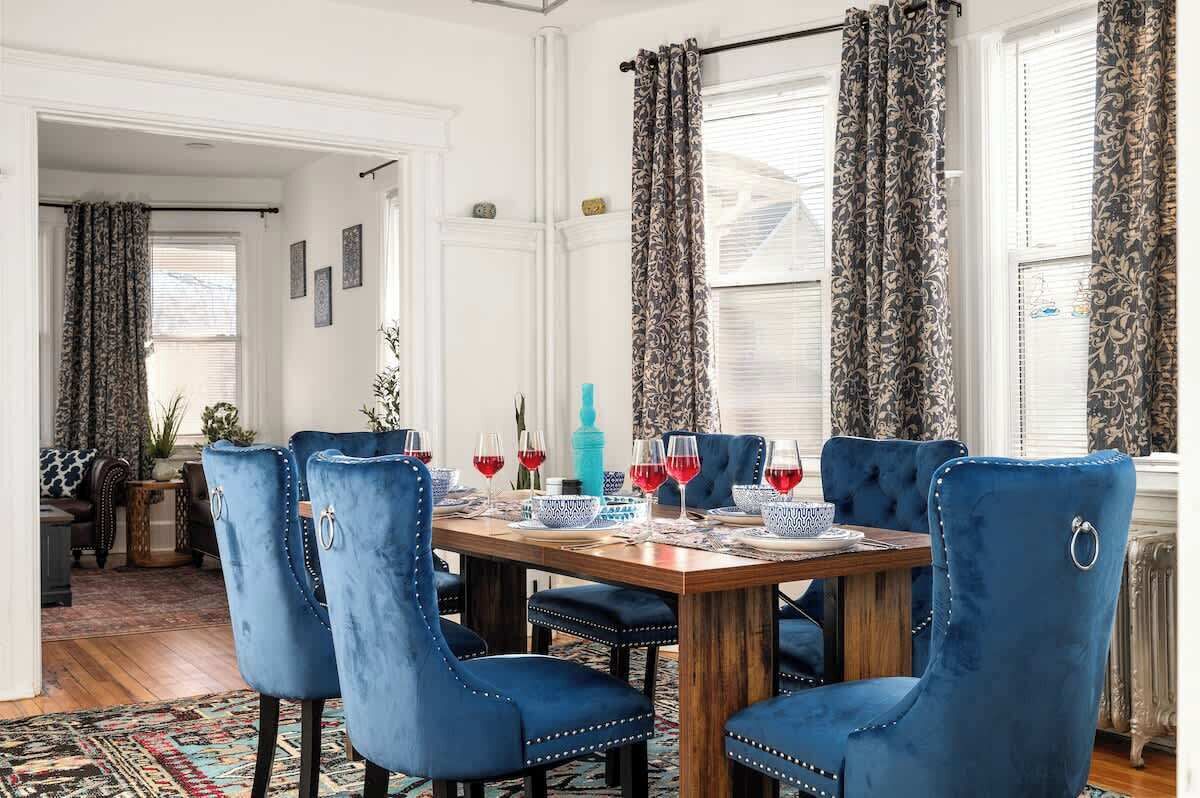 Un coin repas avec une table en bois, des chaises à l’assise rembourrée de tissu bleu et des rideaux à motifs.