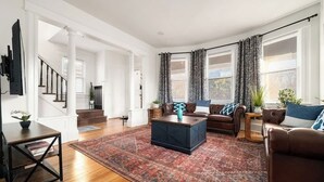 A spacious living room with a large rug, a coffee table, a sofa, and a TV mounted on the wall. - Near NYC & Transit ~ Family Friendly ~ Whole House (New Rochelle)