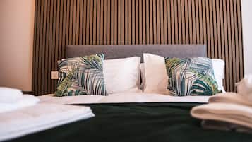 Una cama bien hecha con cabecera gris, almohadas estampadas en blanco y verde, y colcha verde oscuro.